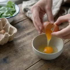 Mani rompono un uovo in una ciotola, mentre altri uova sono in un cartone. Attenzione, le uova fanno male al fegato se consumate in eccesso.