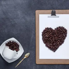 Chicchi di caffè a forma di cuore su una lavagna, a ricordare che il caffè fa bene al cuore. Una tazzina e un cucchiaino dorato completano la scena.
