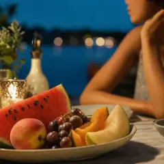 Piatto di frutta fresca con anguria, melone, pesche e uva. Una donna sullo sfondo, forse pensa che la frutta fa ingrassare, ma è deliziosa!