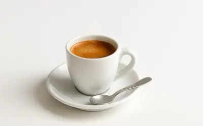 Tazzina di caffè espresso su piattino bianco con cucchiaino. Un piacere quotidiano, ma attenzione al caffè e colesterolo.