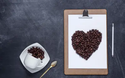 Chicchi di caffè a forma di cuore su una lavagna, a ricordare che il caffè fa bene al cuore. Una tazzina e un cucchiaino dorato completano la scena.