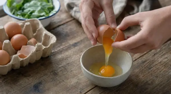 Mani rompono un uovo in una ciotola, mentre altri uova sono in un cartone. Attenzione, le uova fanno male al fegato se consumate in eccesso.