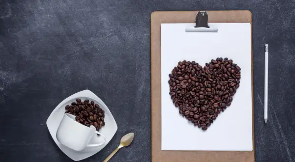 Chicchi di caffè a forma di cuore su una lavagna, a ricordare che il caffè fa bene al cuore. Una tazzina e un cucchiaino dorato completano la scena.
