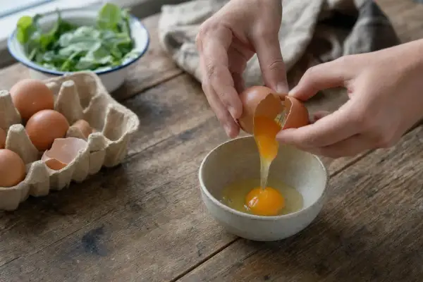 Mani rompono un uovo in una ciotola, mentre altri uova sono in un cartone. Attenzione, le uova fanno male al fegato se consumate in eccesso.